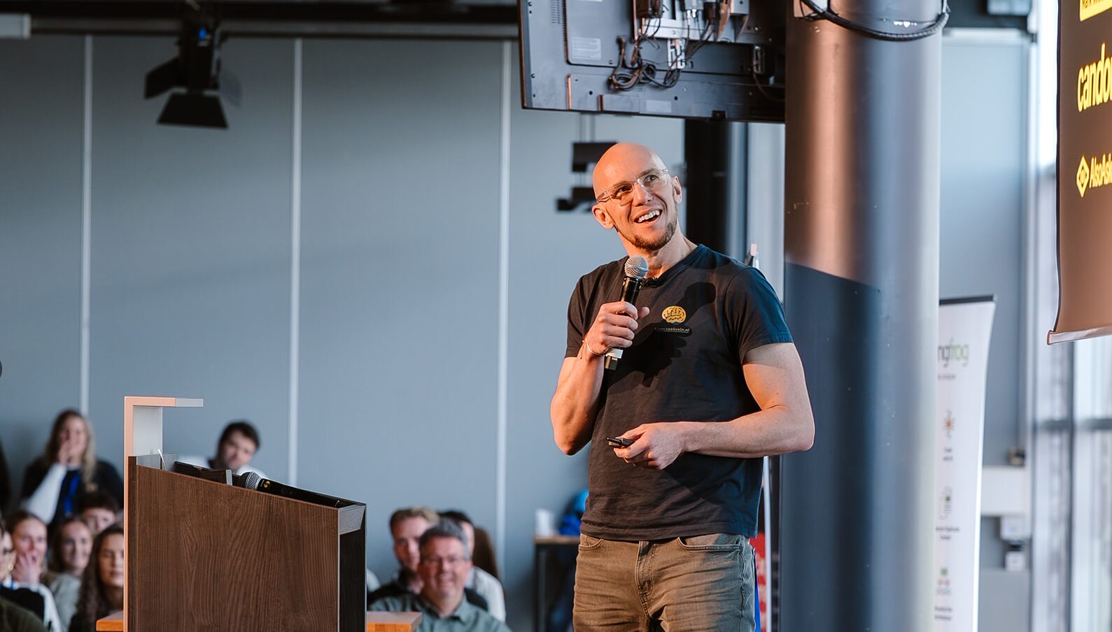 with glasses smiling and speaking into a microphone during a presentation, with an out-of-focus audience seated in the background.